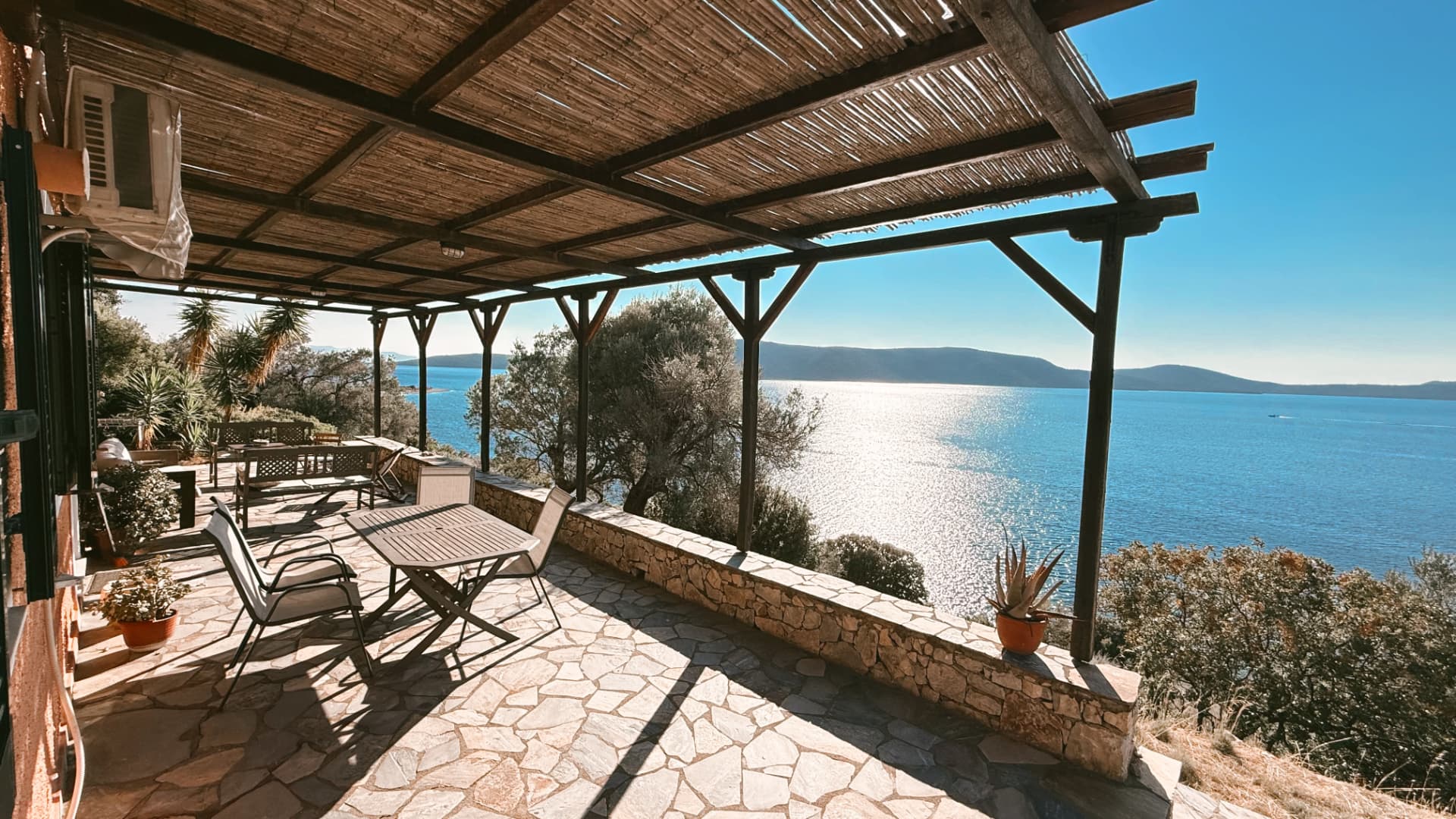 Covered stone terrace with pergola seating overlooking the Aegean at golden hour